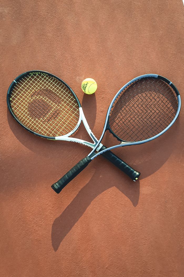 about-us Two tennis rackets and a ball arranged on a clay court, casting shadows.