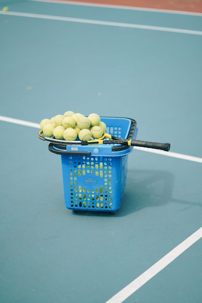 about-us A tennis racket and balls placed on a blue tennis court with a basket, depicting a sporty theme.