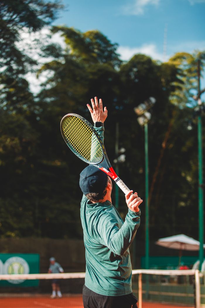 our-story A man in a cap serves a tennis ball during a match on an outdoor court.